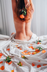 Cropped view of naked woman sitting backside on soft white bedding with orange red roses and petals...