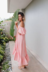 Full-length shot of graceful caucasian girl posing with green plant. Outdoor photo of cute fair-haired lady wears long pink gown.