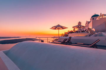 Amazing view over white architecture with colorful sky. Chaise lounge with umbrella under sunset view. Beautiful travel and vacation background, sunset on Santorini island, Greece.