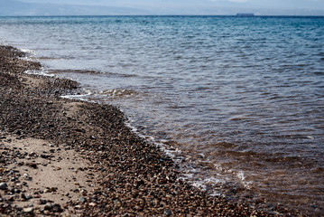View at the beach with dark sand and small stones with blue turquoise sea water. Tropical resort scenic background. Seaside under the beams of raising sun in the morning.