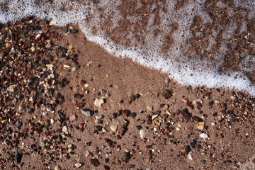 Close-up picture of sandy beach sand with small colorful stones rocks and foamy waves of sea water. Natural texture tropical background.