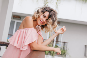 Stylish girl with big eyes posing at balcony. Photo of glad tanned woman in pink attire standing in hotel.