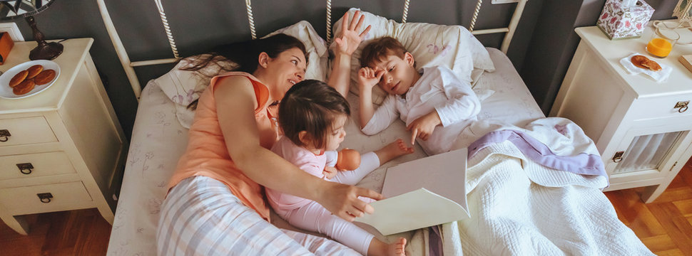 Top view of mother reading story book to her daughter and son kids lying in the bed during virus confinement