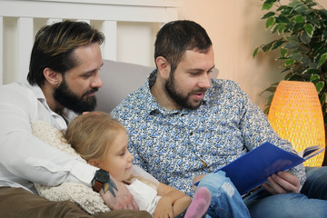 Same Sex Male Couple At Home with daughter. Homosexual couple read a book to their daughter while lying in bed. Stylish gay couple with their child