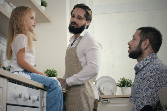 Same Sex Male Couple With Daughter At Home. Homosexual Male Couple Cooking In Kitchen And Talking With Their Daughter