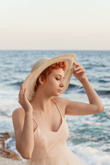 A beautiful, red-haired girl is standing against the sea, wearing a hat on her head, wearing an airy, light dress. Waiting for a lover, the heroine of the novel.