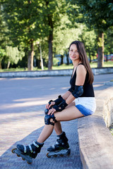 Fototapeta na wymiar Young pretty brunette woman, wearing rollerblades and safety equipment, sitting on a concrete platform, relaxing in a park after ride. Full-length sitting portrait of fit sporty girl in roller skates.