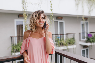 Attractive female model with short wavy hair calling friend from hotel. Photo of lovely young woman in pink blouse talking on phone while standing at balcony.