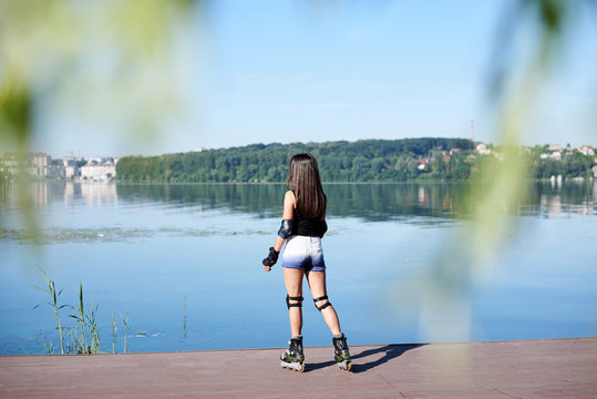 Young pretty brunette woman, with long hair, wearing roller blades with protective equipment, posing, standing on platform by city lake. Full-length portrait of fit slim roller skater girl in summer.