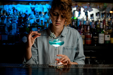 Pretty woman barman decorates glass with blue drink flower branch and look at it.