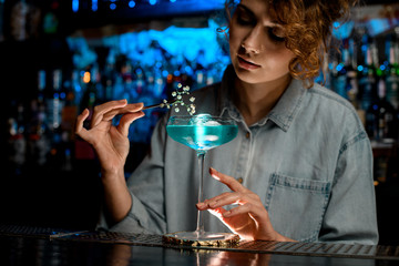Lady bartender decorates glass with blue drink flower branch and look at it.