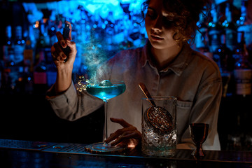 Young woman bartender accurate sprinkles on glass with blue cocktail