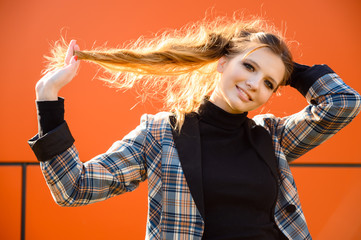 Portrait of a stylish beautiful young girl with long hair with a smile in the park against the background of an orange building wall. Photo taken in sunny weather outdoors in spring.