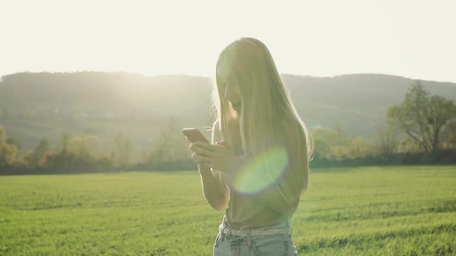Happy young attractive woman use smartphone field landscape cellphone happy mobile girl nature walk sunlight green grass sunset portrait slow motion