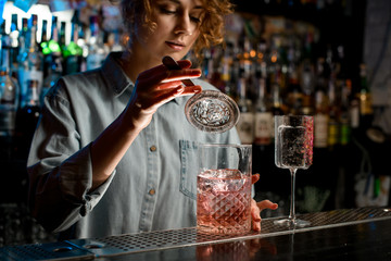 young woman's hand holds strainer over large glass with pink drink