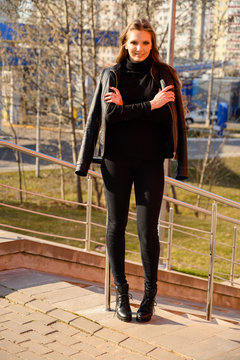 Full-length portrait of a stylish young woman with long hair with a smile in the city on the background of the steps of the building. Photo in sunny weather outdoors in spring.