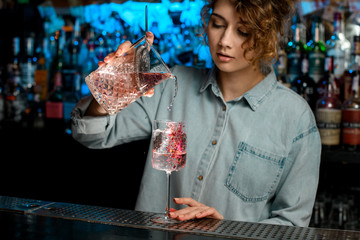 Lady barman pours pink cocktail from large glass into wineglass.