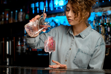 Lady barman carefully pours pink cocktail from large glass into wineglass.