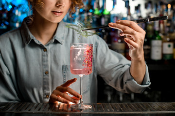 young woman bartender accurate decorates glass with cocktail by piece of green plant.