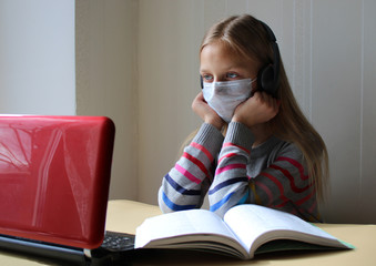 Blonde girl in a medical mask with headphones sits at a table with a computer and a book. Home schooling. The school is closed on quarantine due to the pandemic of Covid-19. Distance learning
