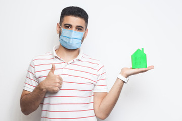 Stay at home, stay safe. Positive man with surgical medical mask holding green paper small house, propose to be at home on quarantine time, thumbs up. Indoor, isolated, studio shot, white background