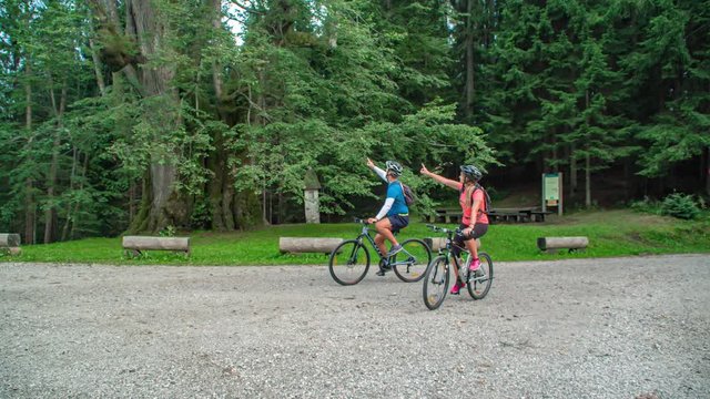 Couple ride bikes on gravel pavement past camera in green forest, slow