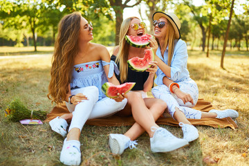 Three beautiful young girls have fun together and eating watermelon  in hot summer day. Friends holding slice of watermelon and posing in the park. Summer concept. 