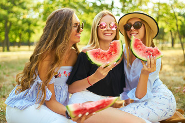 Three beautiful young girls have fun together and eating watermelon  in hot summer day. Friends holding slice of watermelon and posing in the park. Summer concept. 