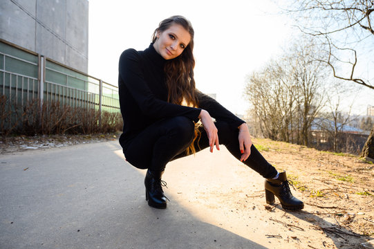 Portrait of a pretty young woman crouched with long hair with a smile on the background of a road in the city. Photo in sunny weather outdoors in spring.