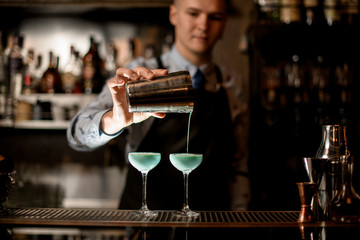 Young barman professionally pours ready-made cocktail from shaker into glass.