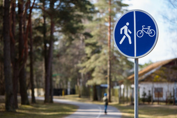 Bicycle and pedestrian shared route road sign over out of focus background of road in park surrounded by forest and houses.
