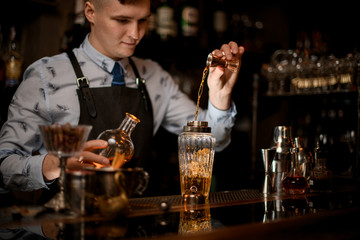 Young handsome bartender carefully pours drink to glassy shaker using beaker.