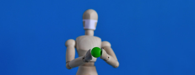 Wooden figure of a mannequin in a medical mask holding a green pill on a blue background. Coronavirus, pandemic and epidemic concept