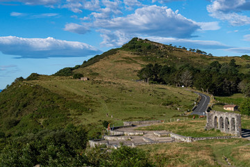 Fototapeta na wymiar View of a hiking trail, old ruins and road on grassy mountain and blue sky with clouds at the background on a sunny summer day. Basque Country, Spain. Camino de Santiago.