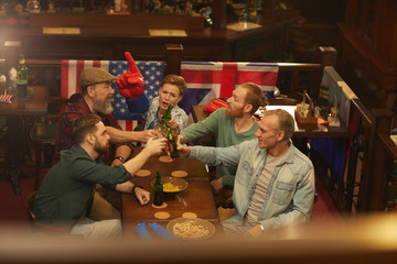 Group of men toasting with glasses of beer while woman cheering for the sport team they sitting at the table in sport bar
