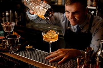 Young smiling bartender pours finished cold cocktail from glassy shaker into glass.