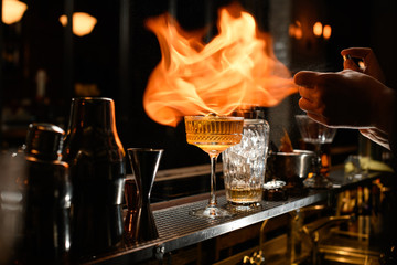 Glass of alcoholic drink and ice stands on bar counter. Bartender makes fire flame over it.