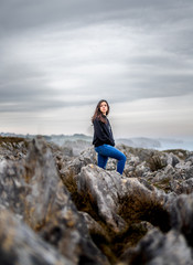Naklejka na ściany i meble Beautiful young girl with black sweater standing on rocks beside cliffs at the coast in a beautiful landscape