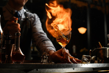 Professional bartender holds glass with cocktail, spraying on it and sets on fire.