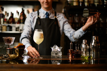 young smiling barman in black apron holds glass with ready-made cocktail