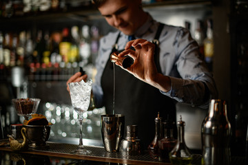 Young barman professionally and carefully pours green drink to steel shaker using beaker.