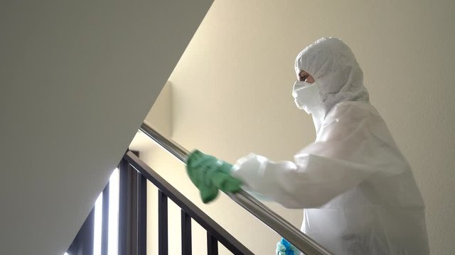 Person protected with a safety suit, mask and gloves to prevent catching a pandemic or virus, cleans and disinfects a doorway of a floor of a house with stairs and elevator