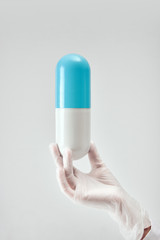 Female hand in white medical glove holding a large plastic blue with white pill vertically against light background