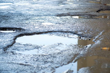 Close up of a road in very bad condition with big potholes full of dirty rain water pools.
