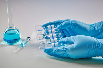 Hands in disposable gloves holding set of ampoules with liquid. Medical flask with blue chemical reagent and syringe isolated on white. Coronavirus