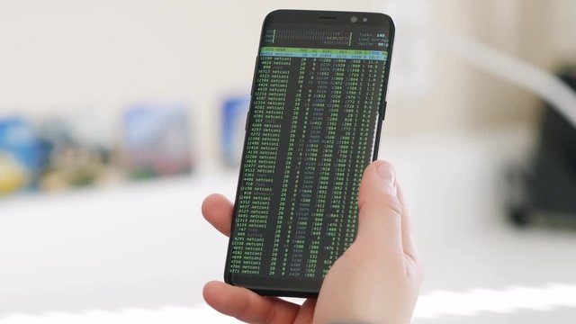 A close up on a hand holding a mobile phone devise that is getting hacked and seeing a security breach via green writings and numbers on the smartphone's screen.