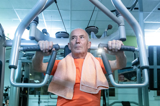Uomo anziano con maglia arancione e asciugamano al collo  si allena in palestra nei macchinari, 