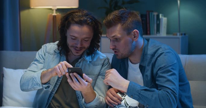Handsome cheerful Caucasian man showing something on smartphone to best friend. Guys football fans putting bets at bookmakers on mobile phone in evening home on sofa. Looking for sport rates online.