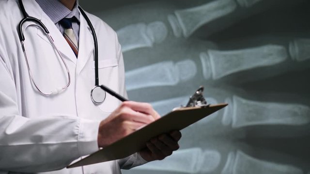 A medical doctor in a white lab coat with a stethoscope taking notes on his notepad on a X-ray scan of hand bones background.Public Domain background element from U.S. Centers for Disease Control and 
