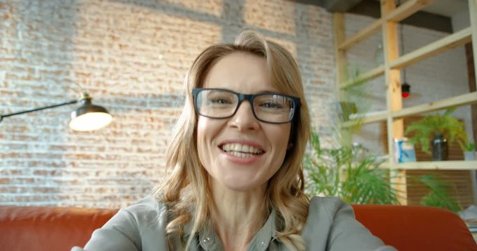POV of happy Caucasian pretty blonde woman video chatting on device at home. Joyful beautiful smiling female model with glasses talking via webcam while sitting in room. Indoor concept. Close up.